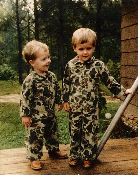 Brison McPhail and his younger brother Payton as little boys in matching camouflage coveralls on a wooden porch