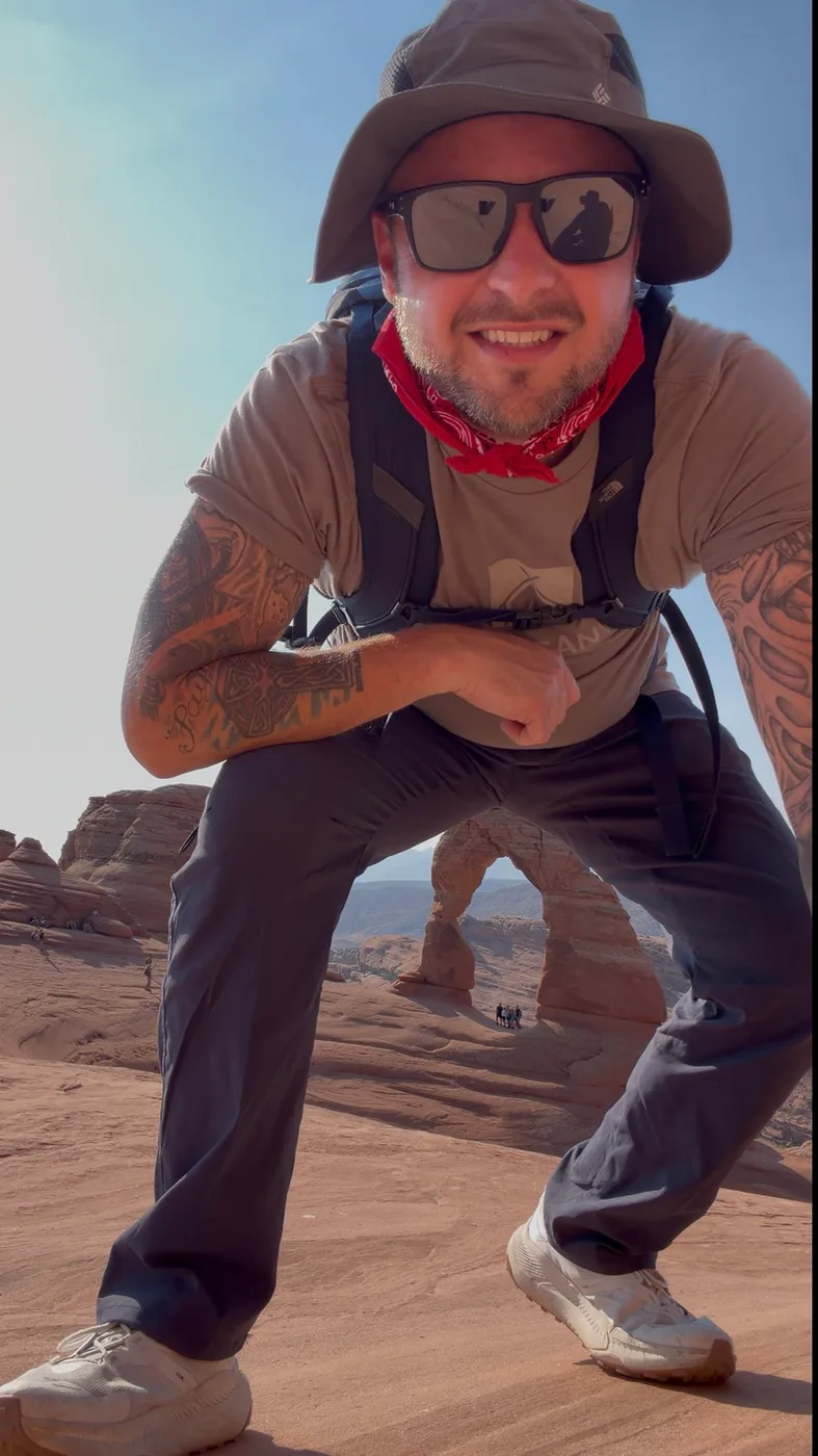 Brison McPhail crouching with Delicate Arch framed behind him