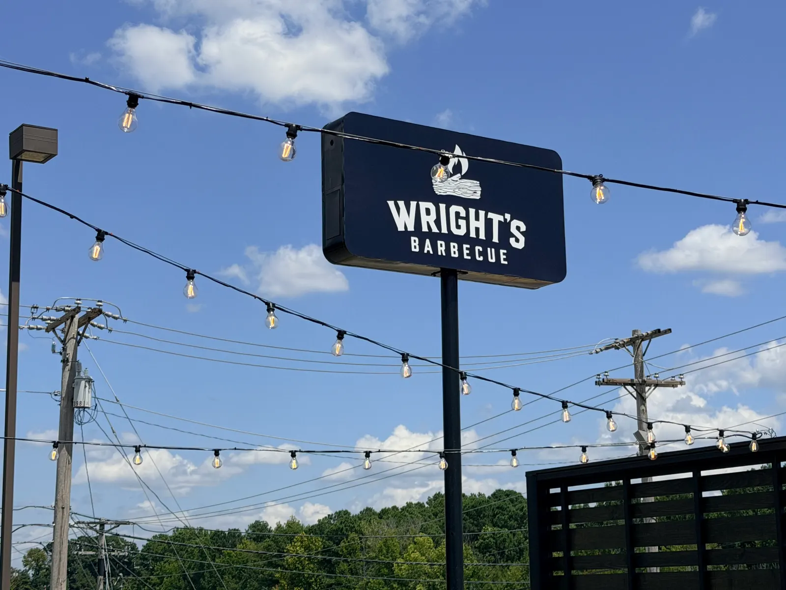 The Wright's Barbecue road sign in Benton, Arkansas — navy sign with a white log-and-flame logo, framed by strings of patio bulbs against a blue sky
